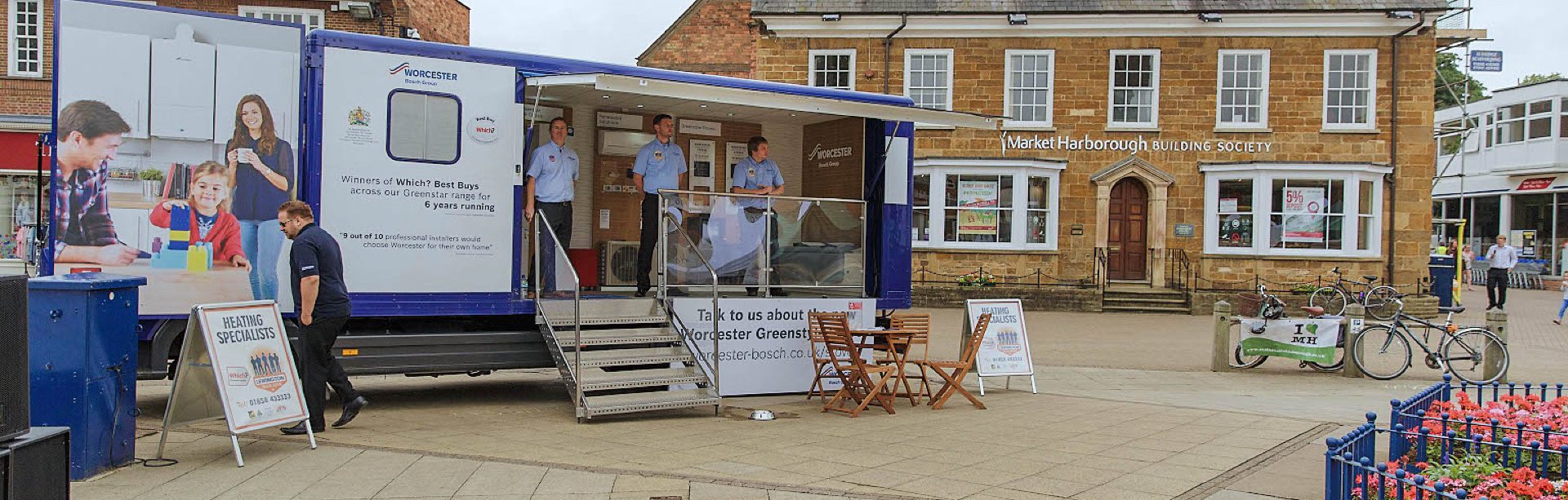 Lewington Heating staff engaging with visitors at a Worcester Bosch exhibition trailer event in a town square, promoting heating services.