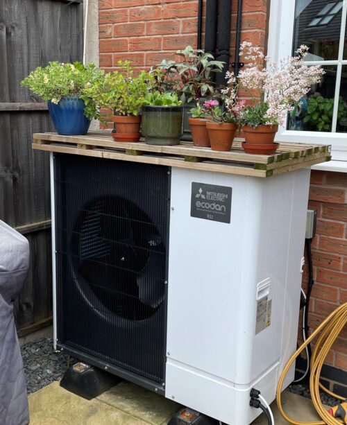 Mitsubishi Electric Ecodan heat pump unit with various potted plants on a wooden pallet cover outside a brick house.