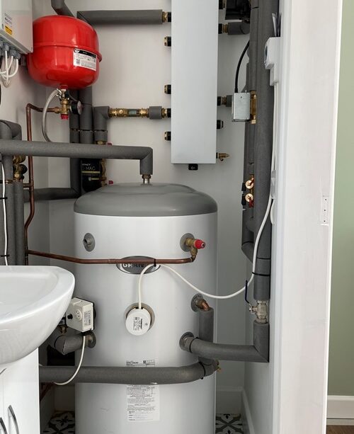 Water heater system with gray insulated pipes a red expansion tank and a white sink in a small utility space.